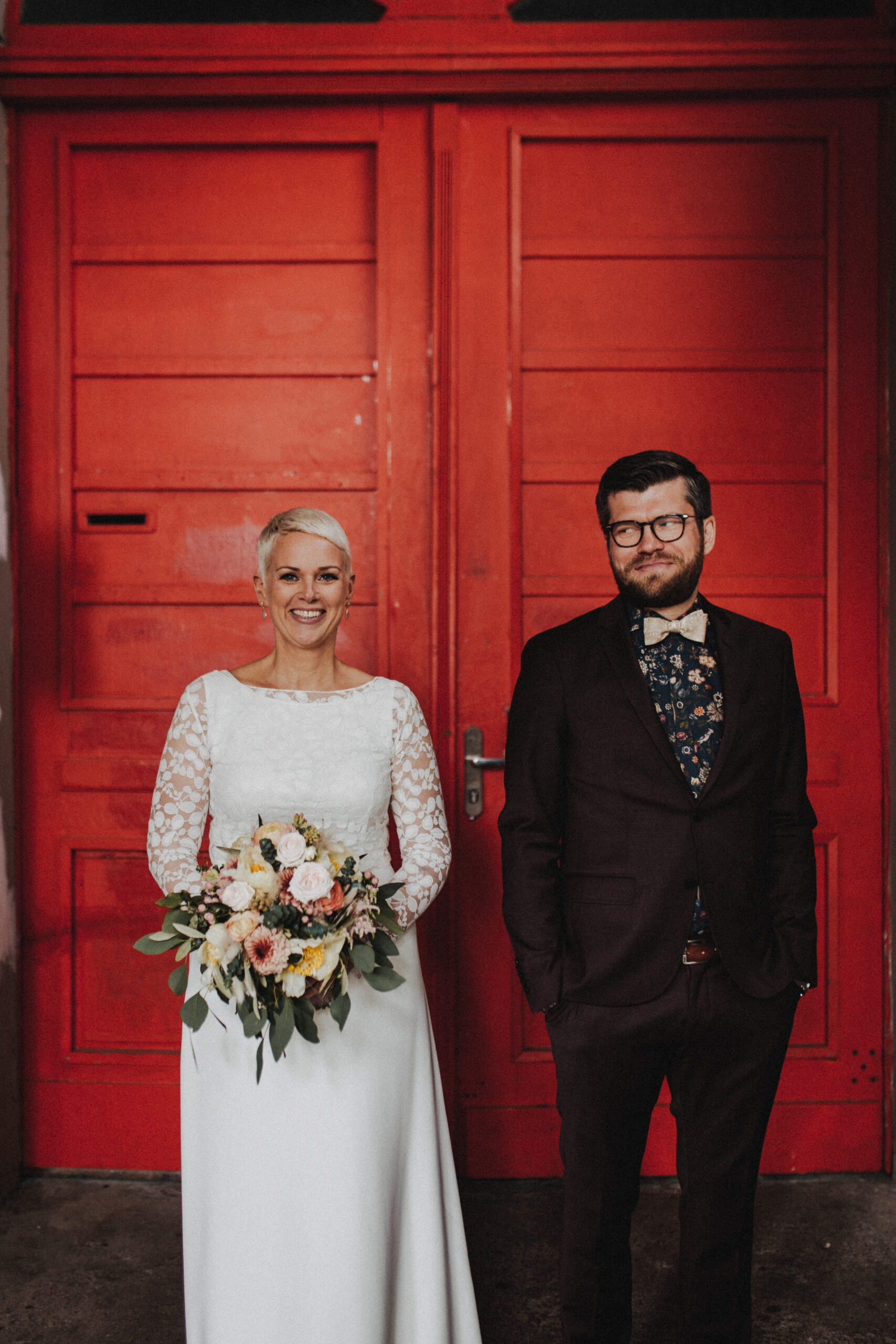Ein Hochzeitspaar vor einer Tür. Die Braut hält einen Strauß. Der Bräutigam schaut die Braut an und lächelt.