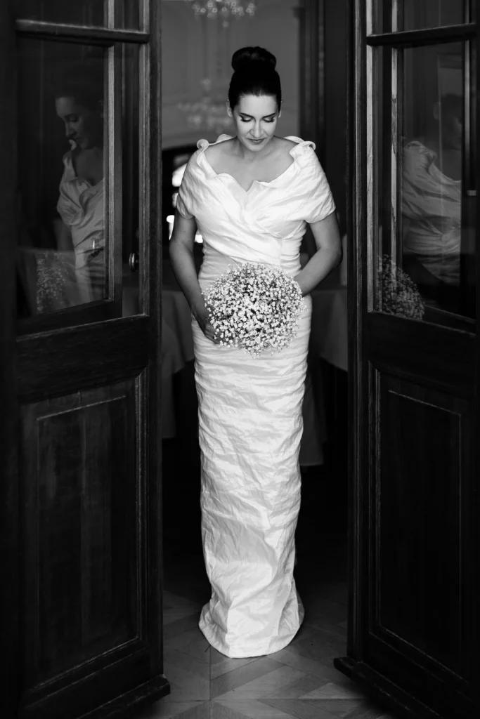 Das Bild zeigt eine Braut, die in der Mitte von Flügeltüren. Sie hält einen Brautstrauß, hat den Blick nach unten gerichtet und ist im Begriff durch die Tür zu gehen.