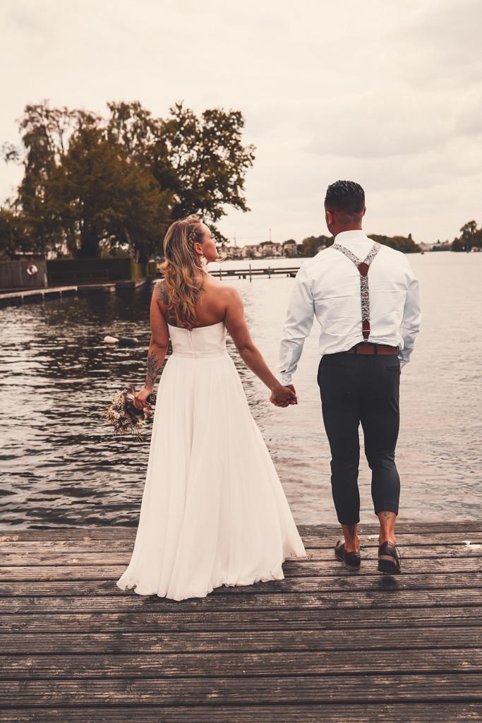 Ein Hochzeitspaar von hinten abgebildet. Es steht, die Hände ineinander fassend, vor dem Wasser mit dem Rücken zur Kamera. Marie-Jo Wieske Punkt d e