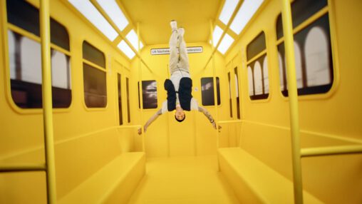 Foto eines Menschen, der in einer durchweg gelben U-Bahn ist. Der Mensch steht kopfüber an der Decke des Wagons und breitet seine Arme aus.